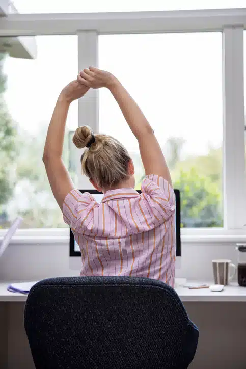 Rear View Of Woman Working From Home On Computer In Home Office Stretching At Desk Rear View Of Woman Working From Home On Computer In Home Office Stretching At Desk